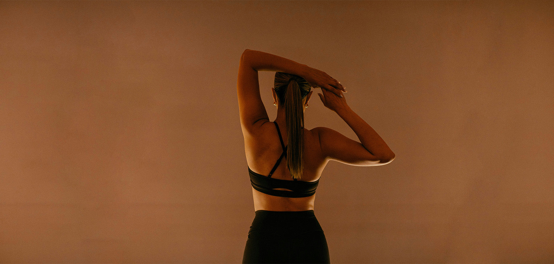 Back view studio shot of woman wearing black cross strap cutout sports bra and high waist leggings, warm neutral tone for Duska yoga fitness activewear collection.