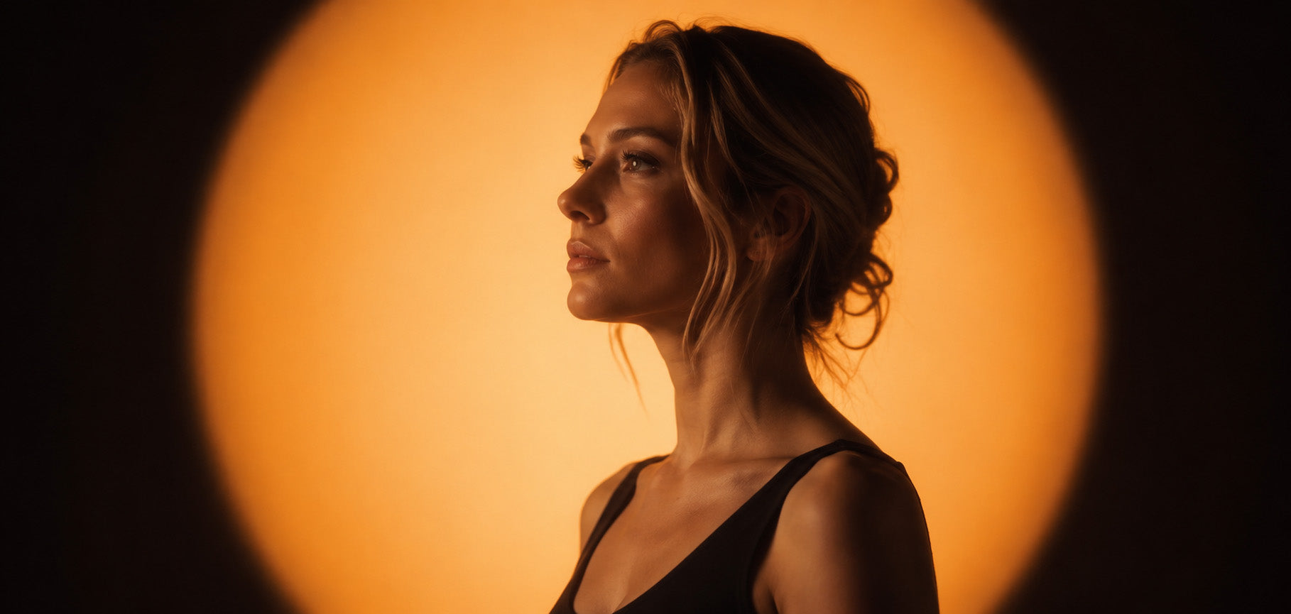 Elegant female side profile portrait under warm circular spotlight, minimalist moody aesthetic homepage hero image for Duska brand, premium lifestyle visual design.