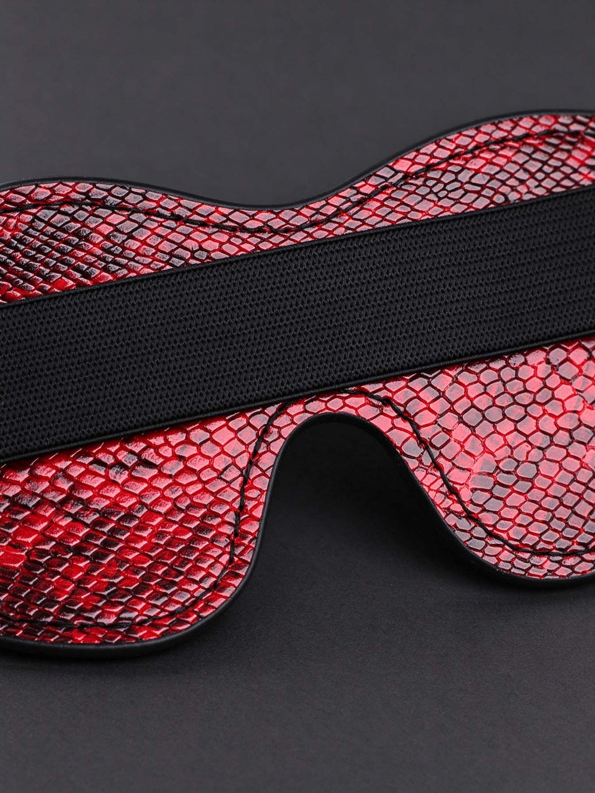 close up detail shot of leather eye mask red snake print faux leather surface black elastic band fine stitching dark grey background sleep accessory