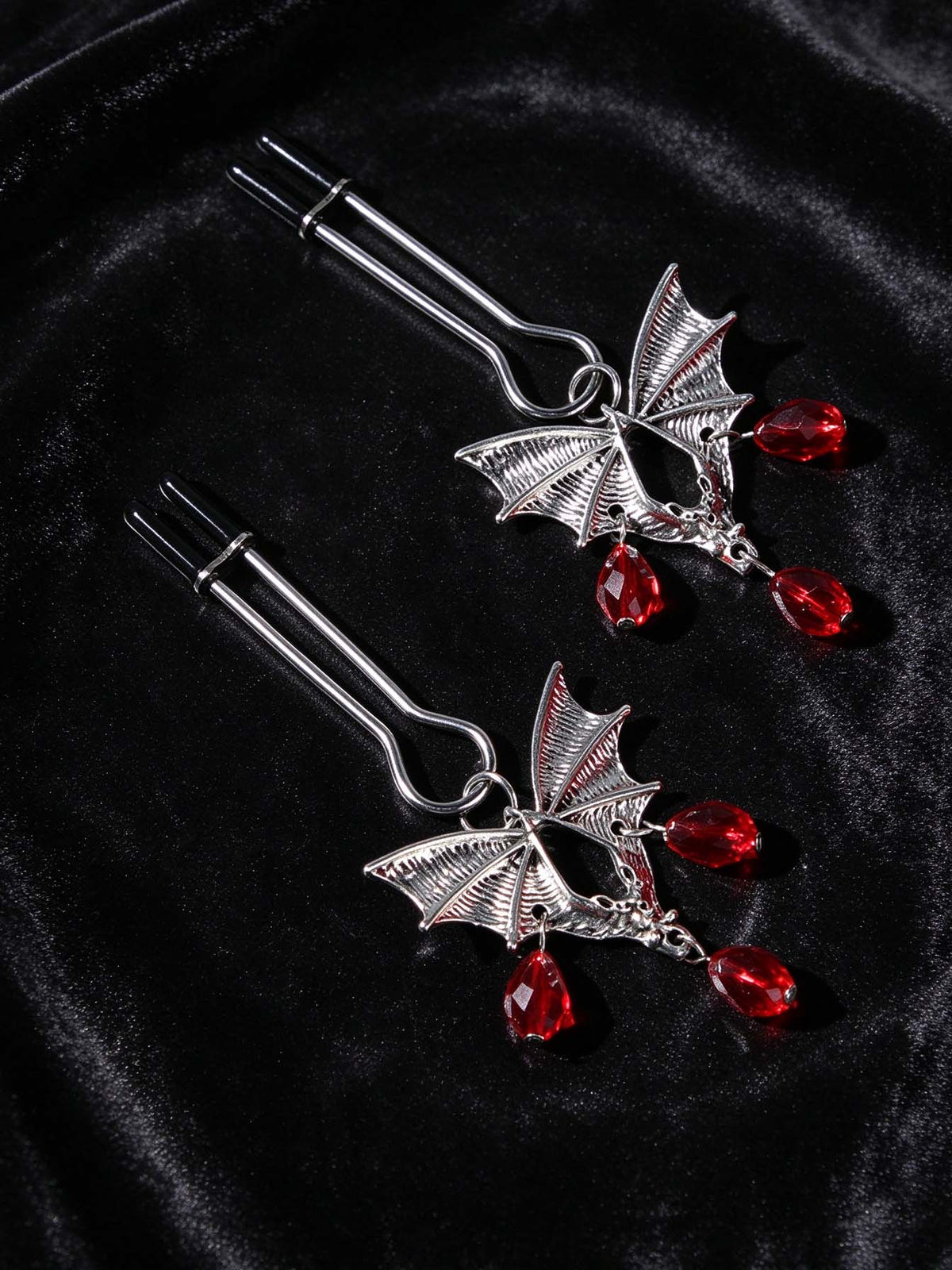 pair of gothic bat nipple clamps antique silver bat pendants with teardrop red crystal beads black rubber tips placed on wrinkled black velvet goth decorative fashion jewelry