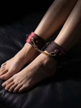 Thermochromic leather ankle cuffs worn on both feet, dark brown leather revealing red pink heat reactive tones, gold D rings and chain, moody low key shot