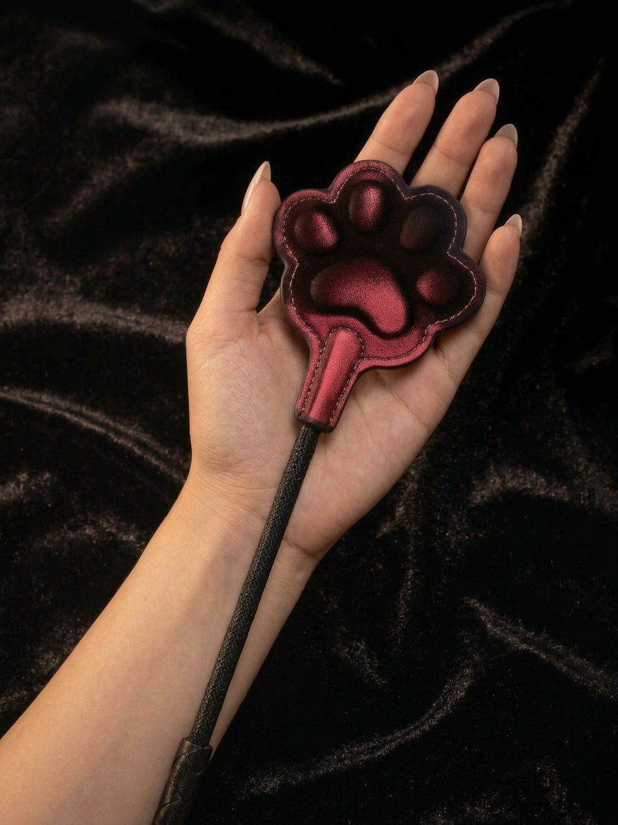 Hand holding thermochromic leather paw paddle, dark red gradient paw shaped pad with black braided handle and wrist strap loop